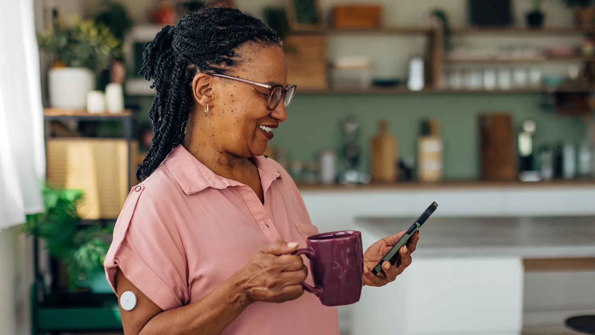 A woman with a blood sugar monitor attached to her arm drinks coffee and looks at her phone.