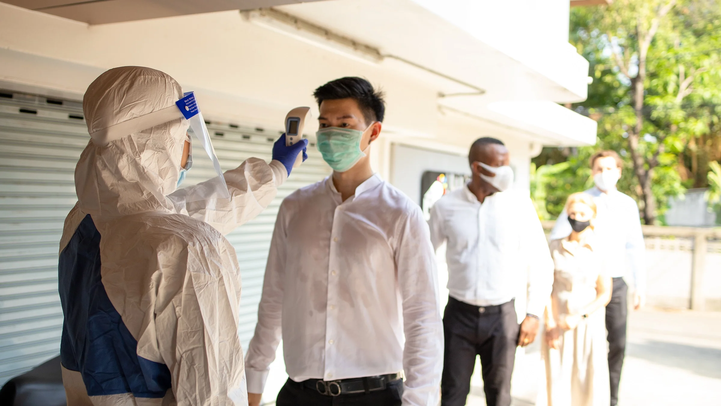 Socially distanced line to enter a building. A person in full PPE gear is checking temperatures as people enter. There four people in line waiting outside.