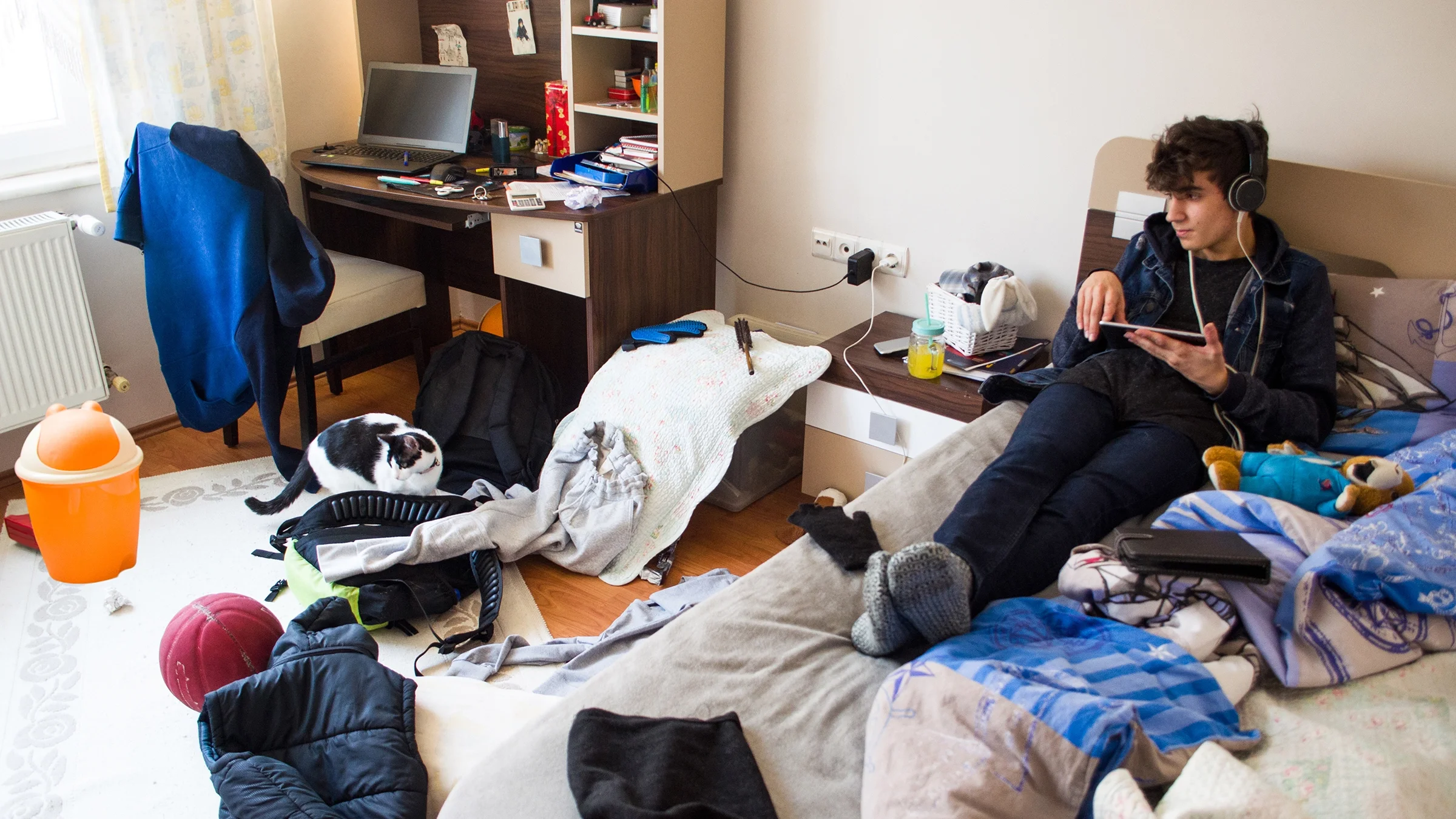 A teenage boy uses a tablet while sitting on a bed in a messy bedroom.