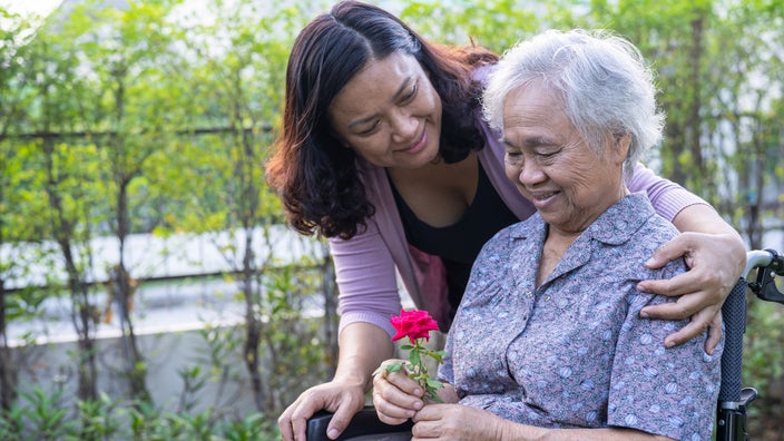 An elderly woman in wheelchair holding a flower and being hugged by her daughter.