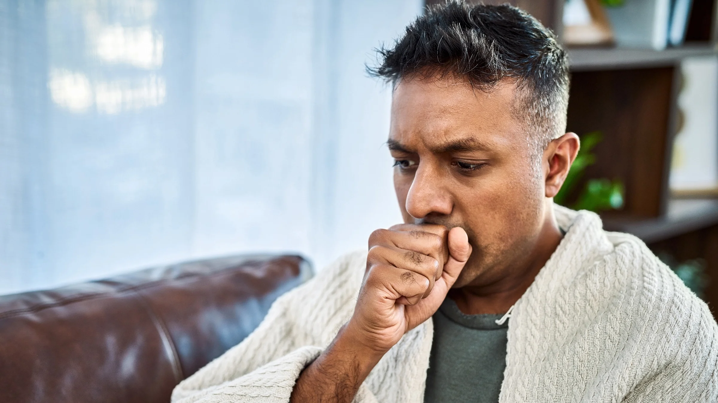 Portrait of a man coughing into his fist. He is wrapped in a blanket and sitting on the couch.