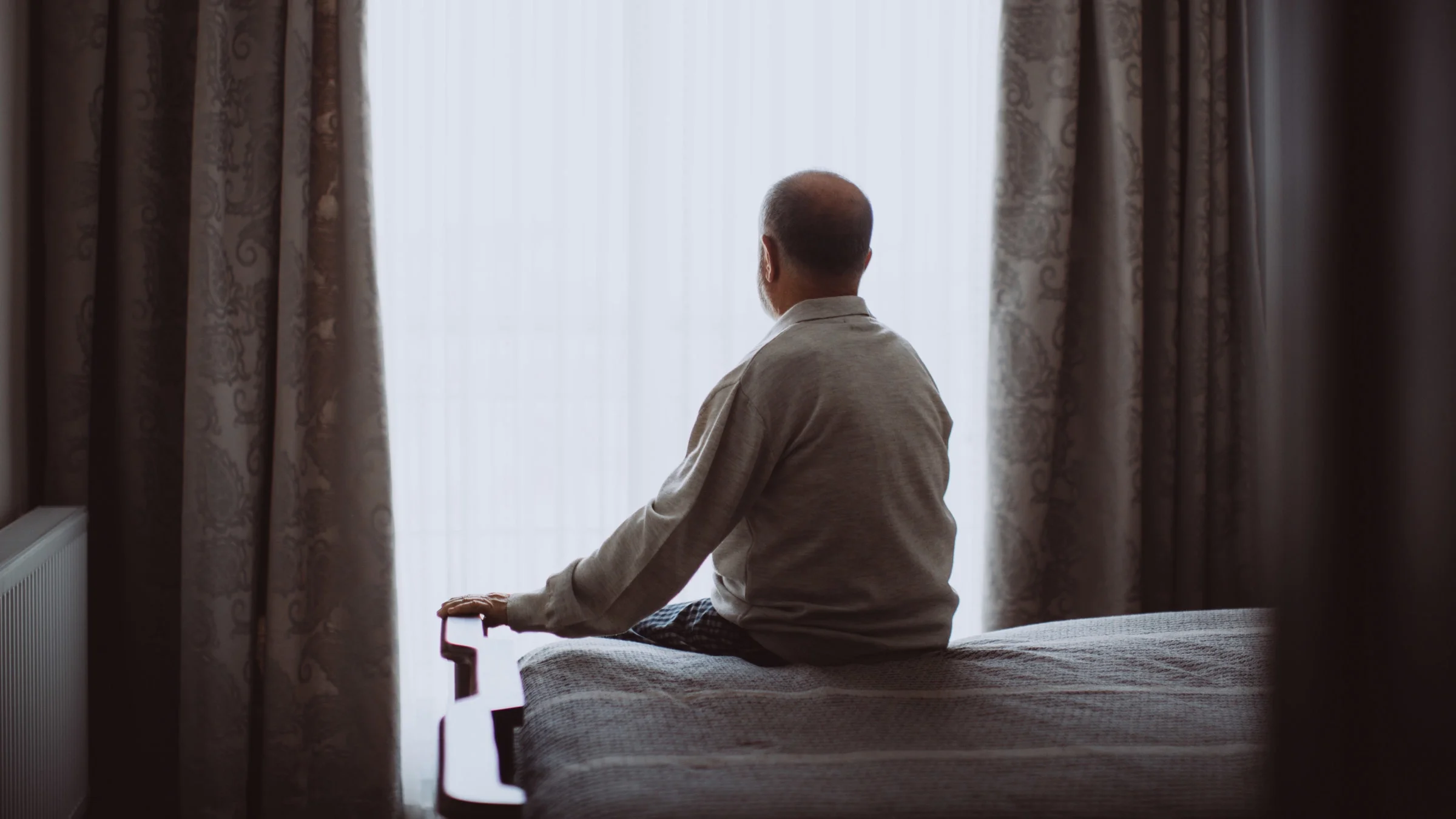 A back shot of someone sitting on their bed, looking outside.