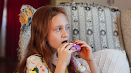 Young girl using a purple inhaler at home.
cometary/E+ via Getty Images