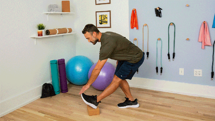 Profile view of man touching his toes while demonstrating standing hamstring stretch with one leg on a yoga block.