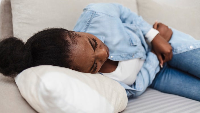 Woman curled up on the couch with stomach pain. She is grasping at her abdomen with her arms.