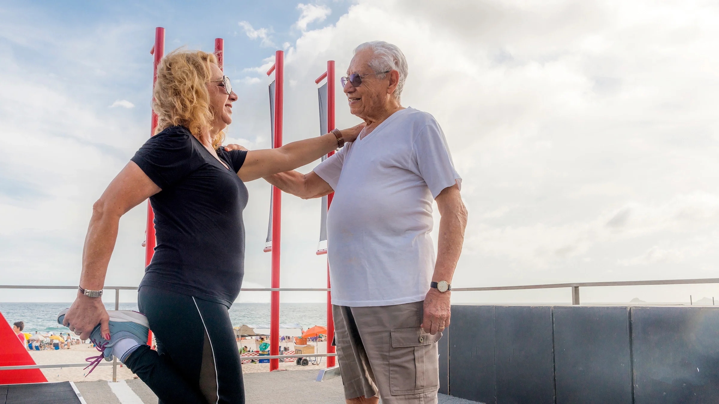 A woman leans on a man's shoulder while doing a standing quadricep stretch.