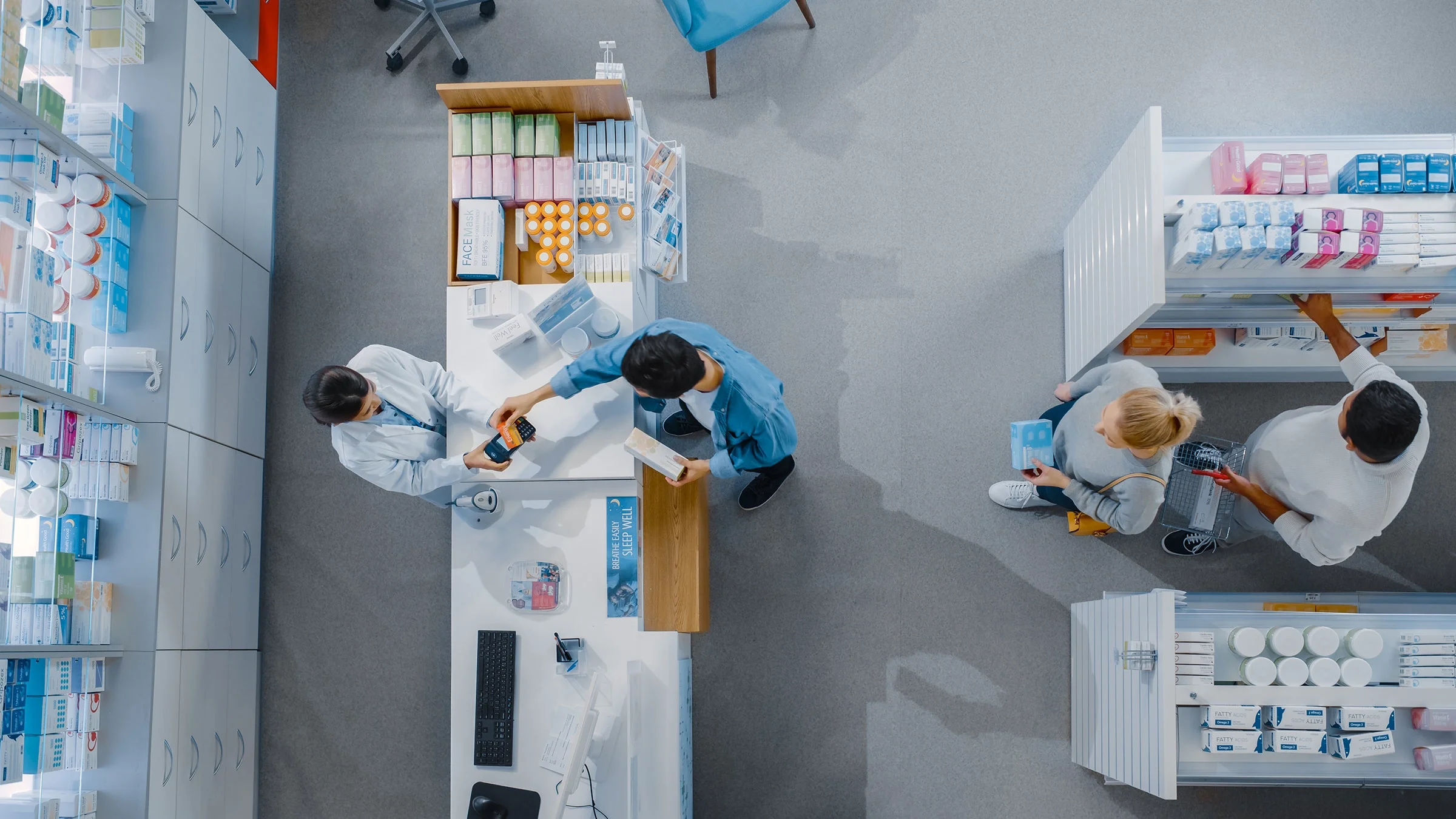 Overhead view of pharmacy checkout counter. A pharmacist is helping out a customer at the counter, while two other people wait in line.