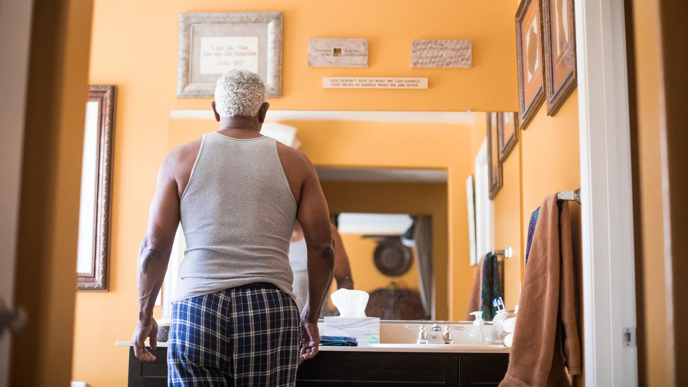 Senior man getting ready in the bathroom mirror. We are looking at his back through the doorway.