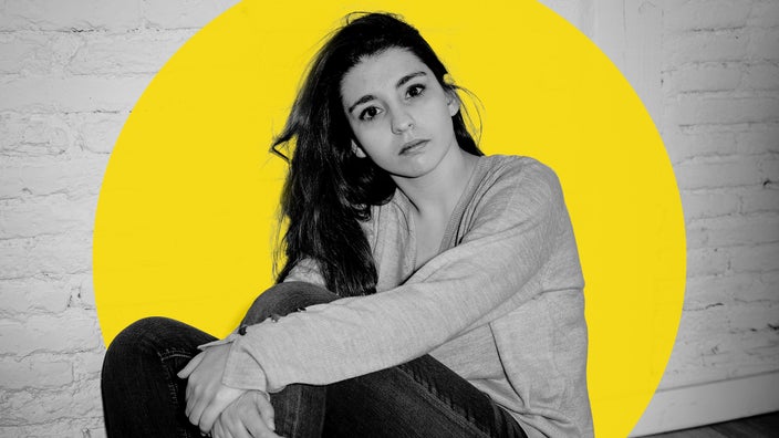 Black and white photo of a woman sitting on the ground. There is a large yellow graphic circle added behind her. She also looks stoic and distant.