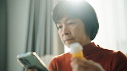 A woman looks at her phone while holding a pill bottle. 
\pocketlight/iStock via Getty Images Plus 