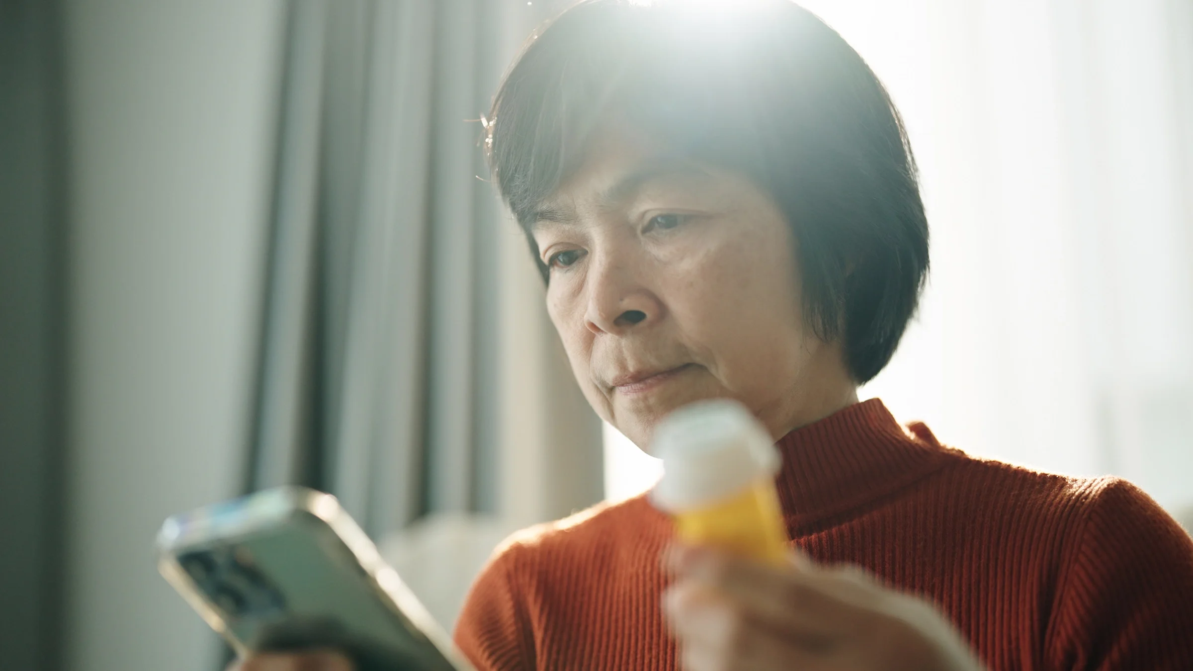 A woman looks at her phone while holding a pill bottle. 