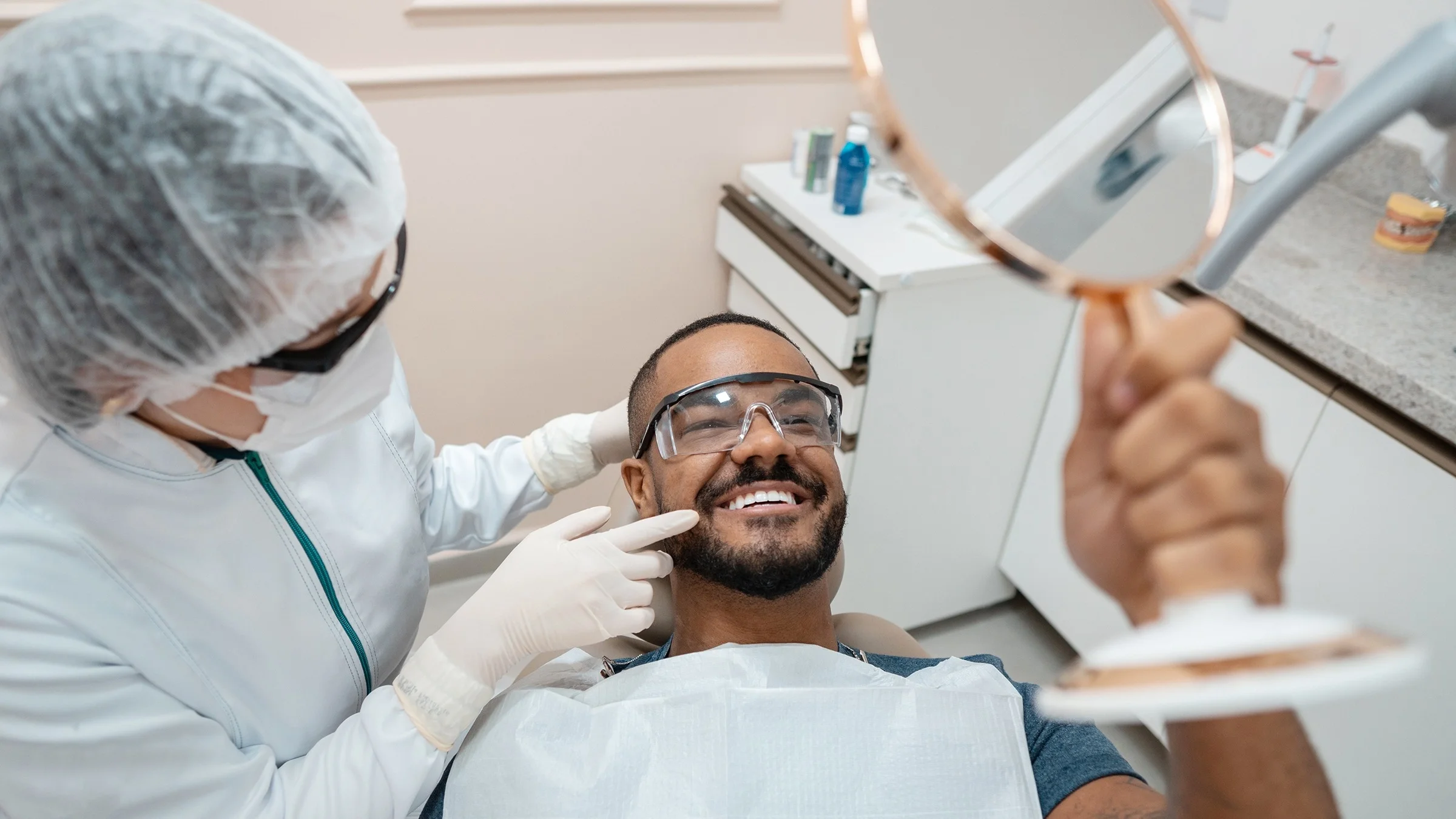 Patient at a dentist visit. He is using a hand held mirror to look at his teeth while the dentist points something out to him.