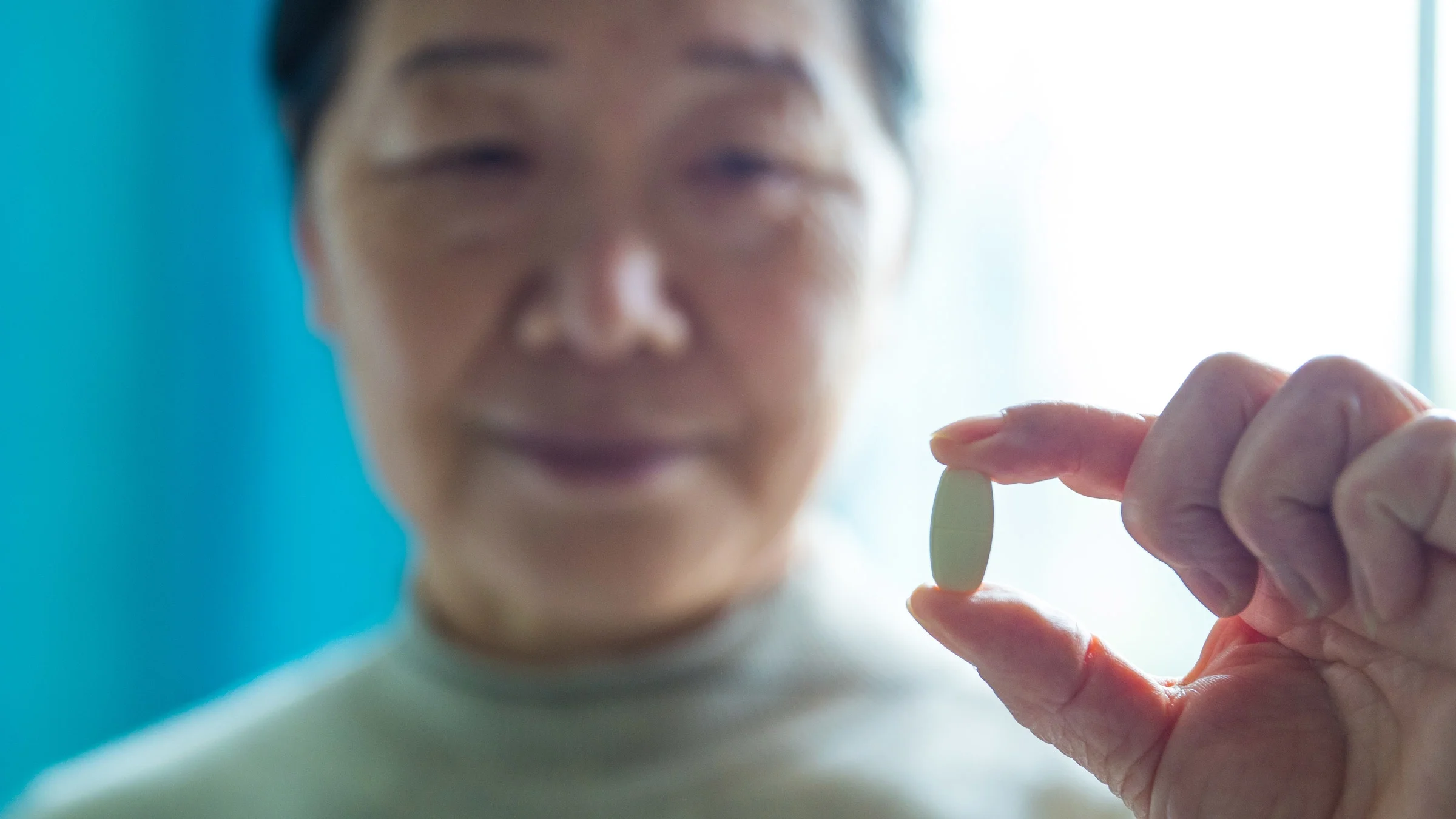 Close-up of a senior woman holding an oblong pill in-between her fingers, the focus is on the pill with her out of focus in the background.