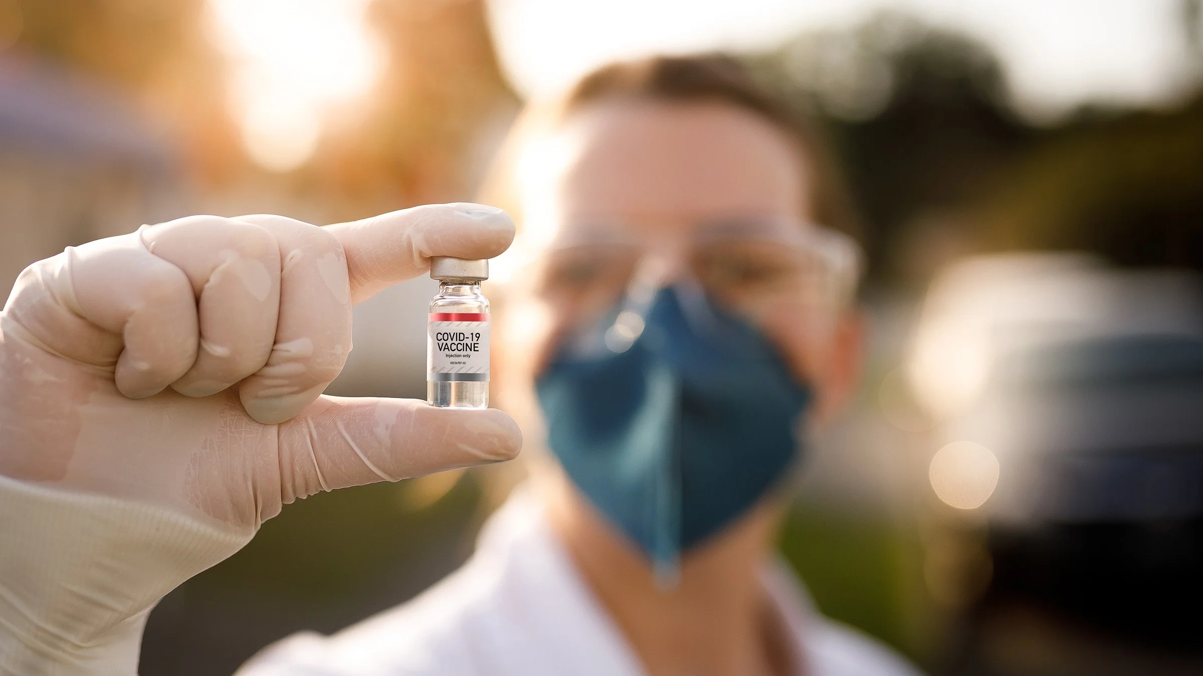Doctor in a dark blue face mask and protective glasses holding up a COVID-19 vaccine vial. The vial is in focus and the doctor is blurry in the background.