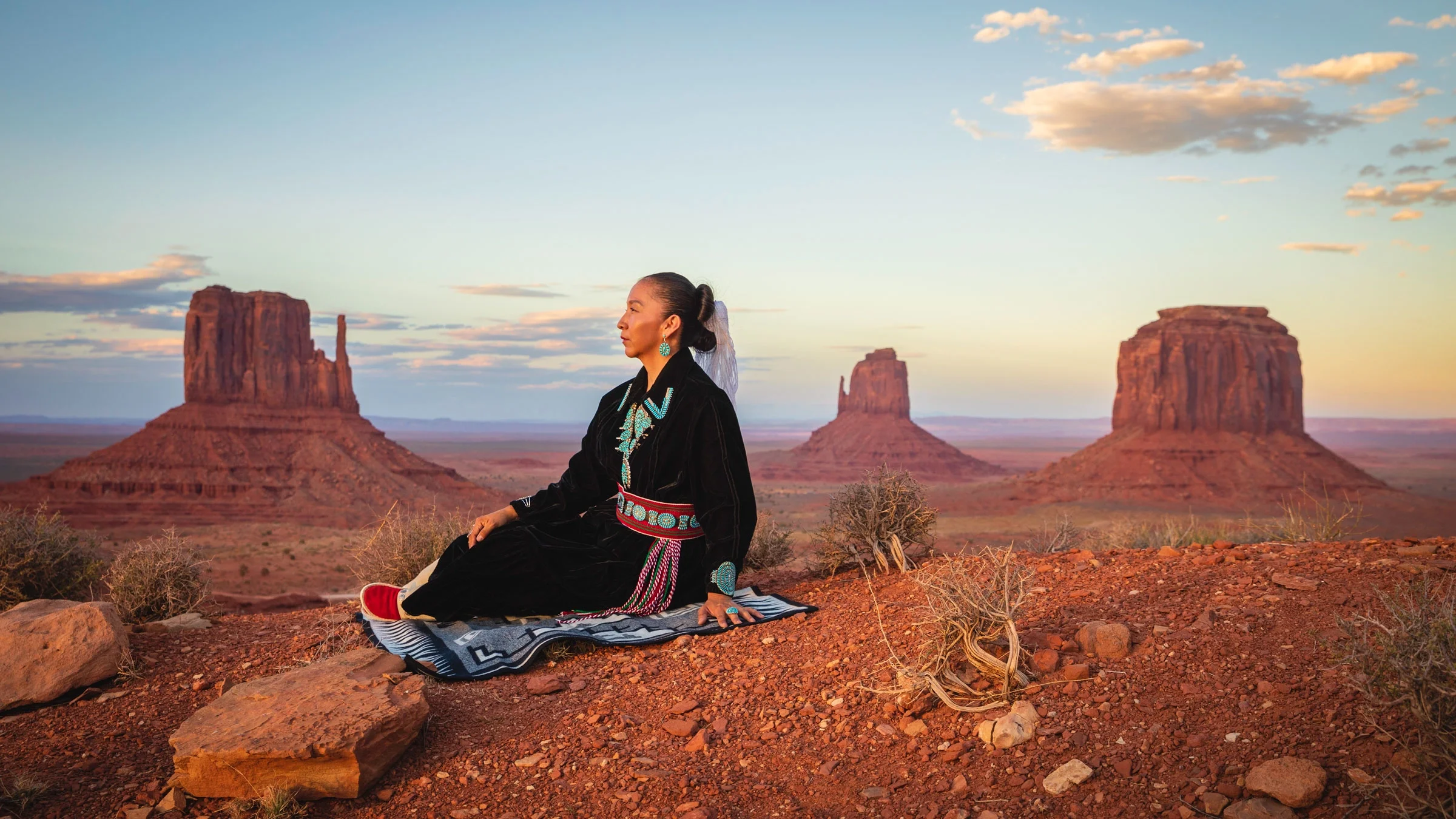 A Native American woman dressed in traditional Navajo clothing poses in the landscape of Monument Valley.