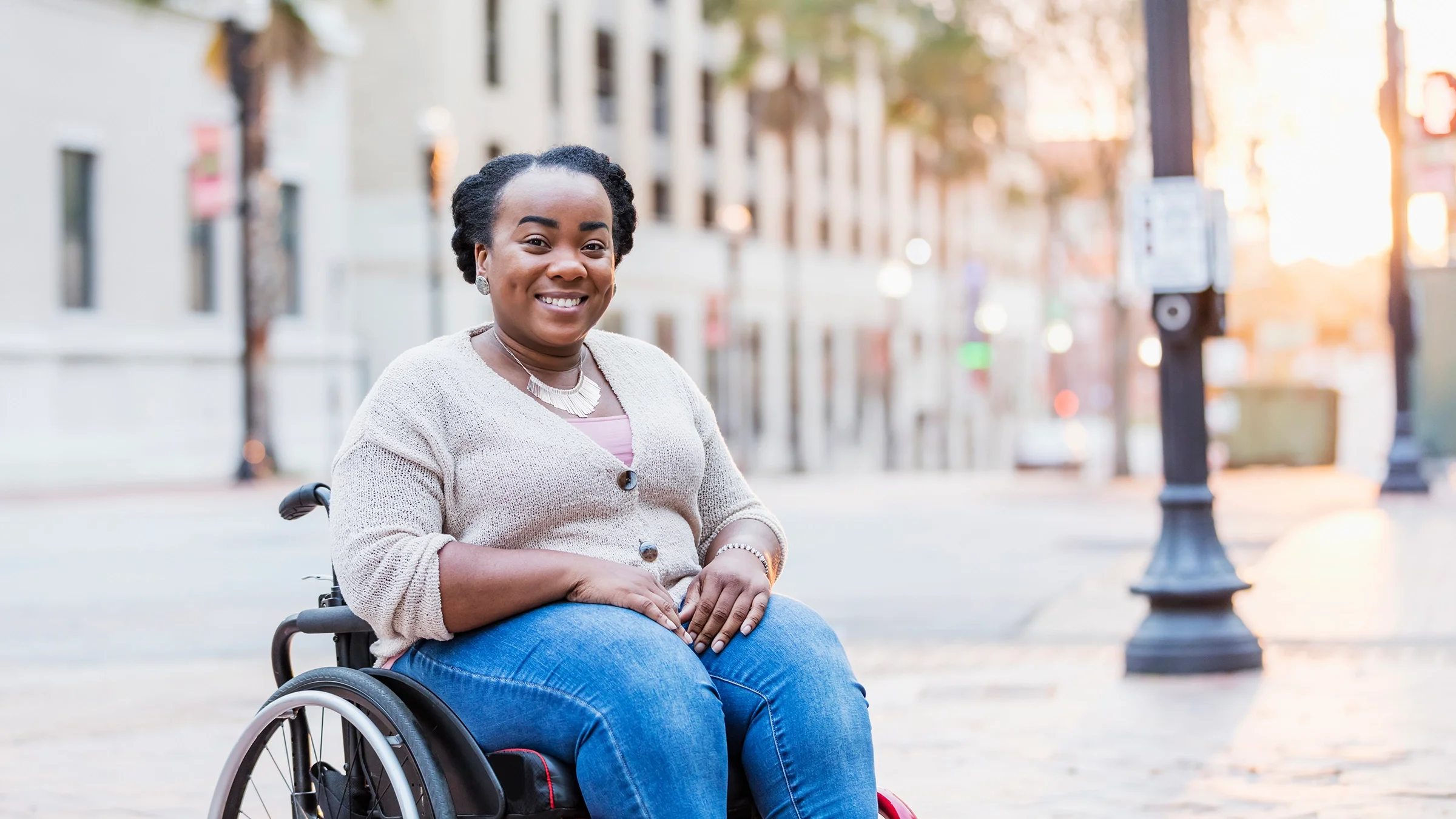 Portrait of a young Black woman in a wheelchair. She is on the sidewalk of a not busy street.