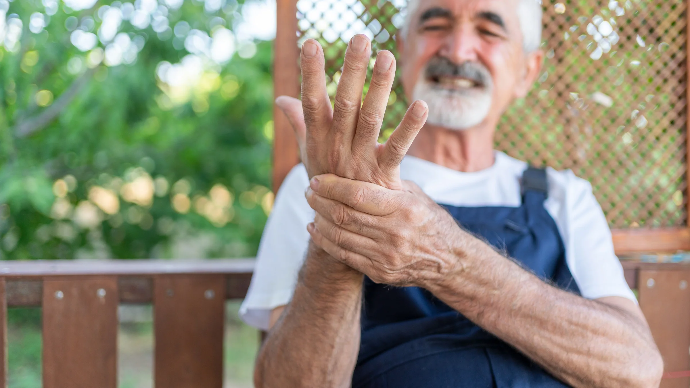 An older adult is focusing on his arthritis in his wrist. He is wearing overalls outside.