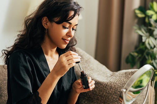 Young woman sitting on the couch in front of a small mirror applying a face serum from the bottle.