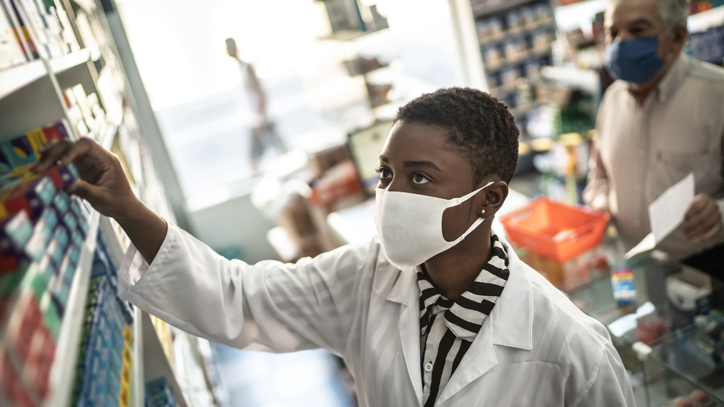 Pharmacist reaching for medication on shelf wearing a white face mask. The patient is in the background blurry standing at the pharmacy counter.