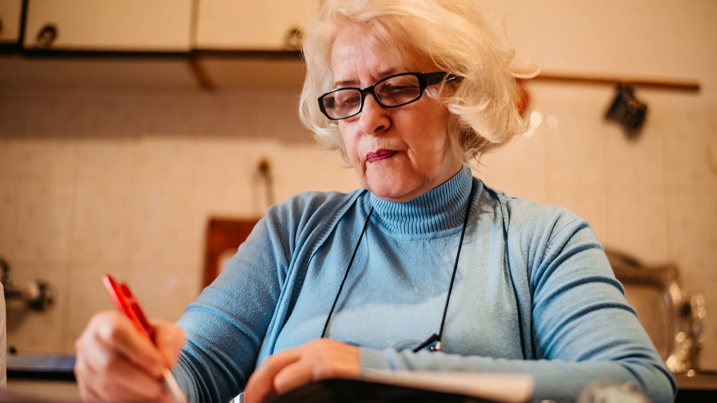 Elderly woman with short white hair writing a letter with a red pen. She is in her kitchen at the dining table.