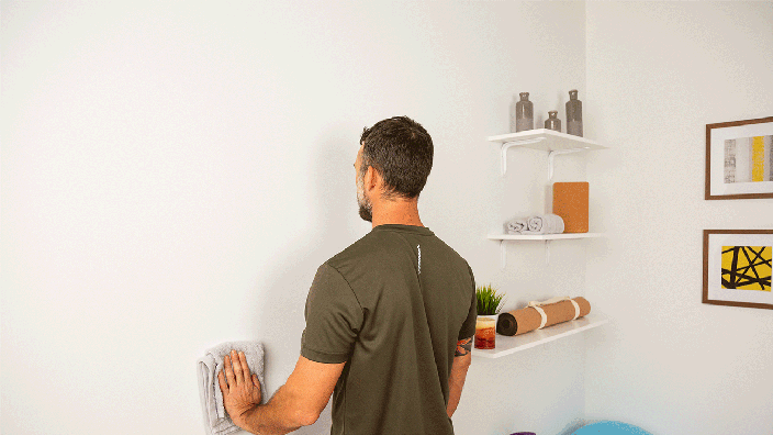A man, viewed from the back, demonstrates the wall slide exercise, sliding a towel up and down a wall. 