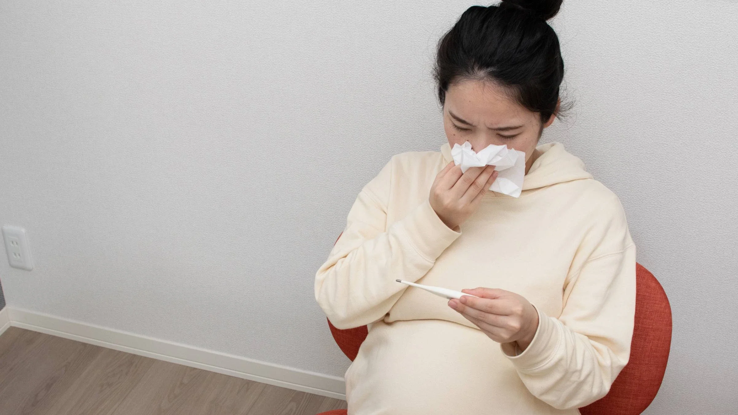 A pregnant person blowing their nose and checking their temperature.