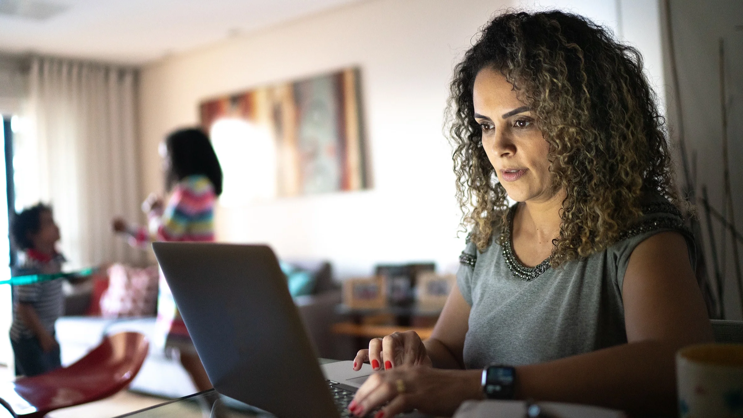 Woman working on her laptop at home with her kids playing in the background in the living room.