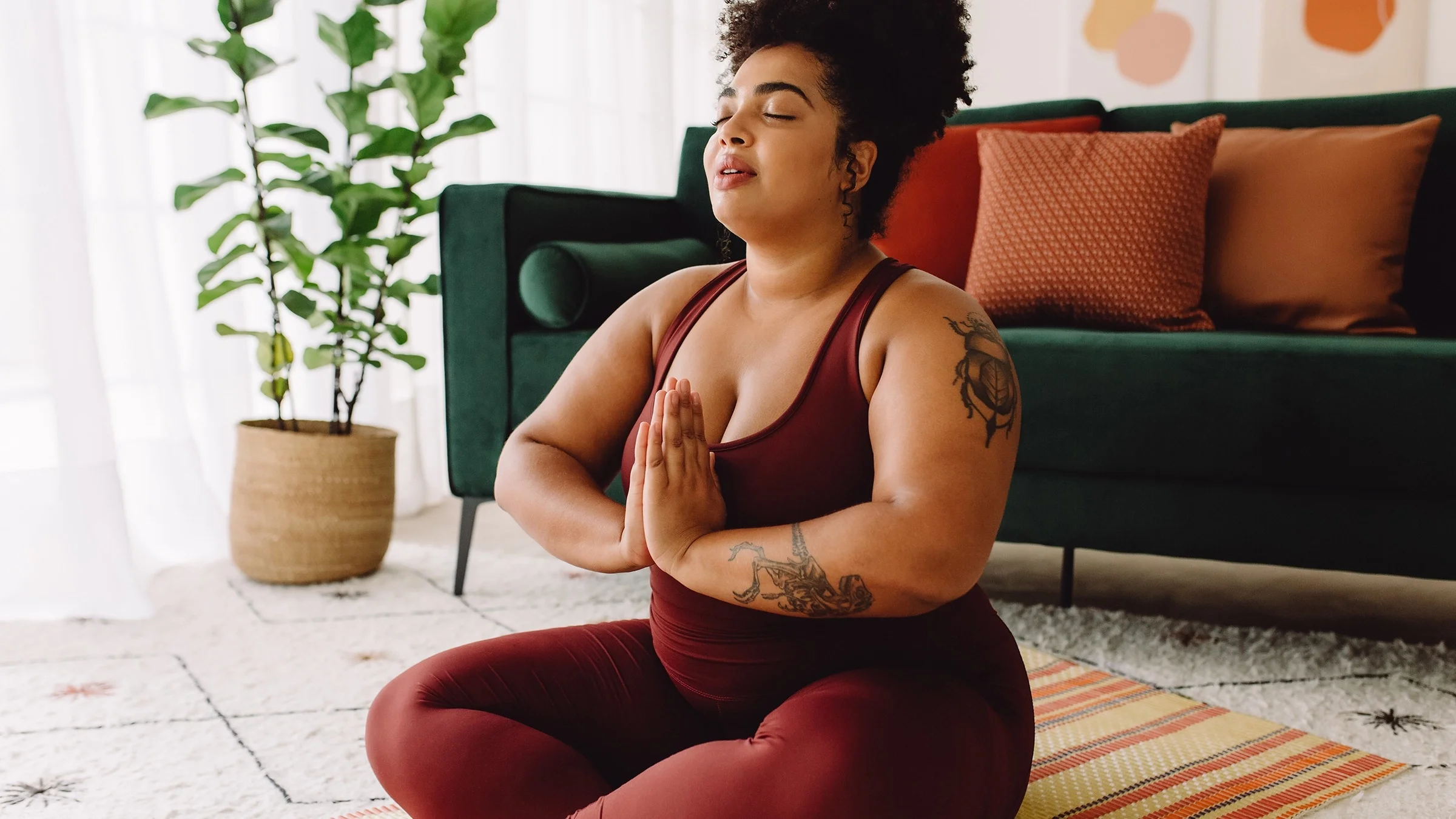 Young Black woman practicing yoga in her living room. She has her hair up high on top of her head and is wearing a dark red workout outfit.