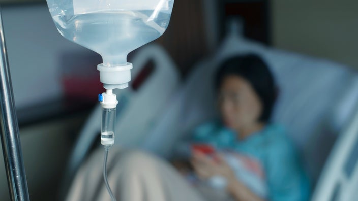 Close-up of an IV saline bag. You can see the patient in the background but blurry on her phone in the hospital bed.