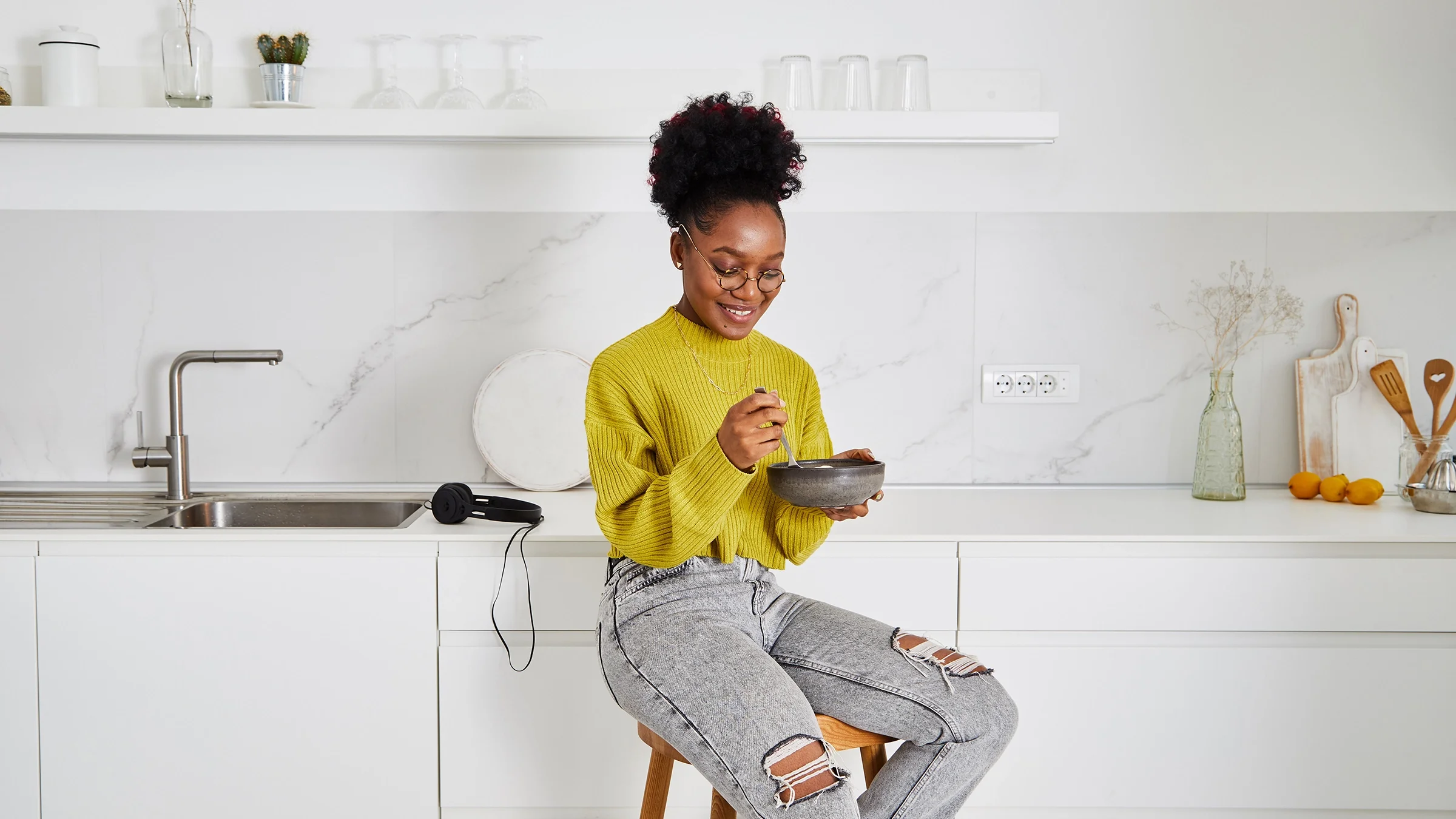 A woman eats breakfast from a bowl while sitting in her kitchen.