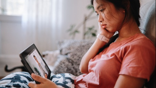 Close-up of a woman on her tablet on a telehealth call with her doctor. The woman is at home on her couch with a blanket on her lap with a sad look on her face.