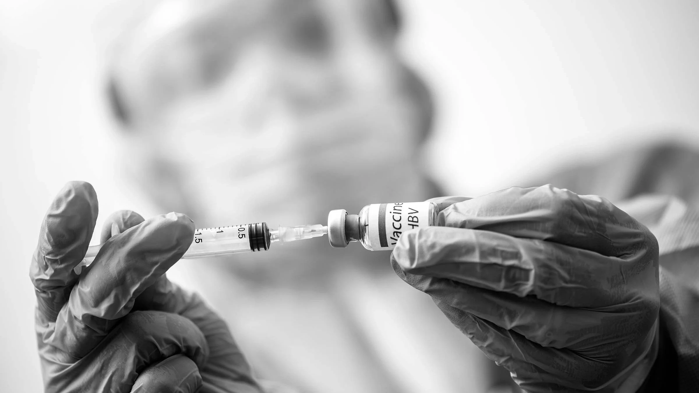 Black and white close-up of a doctor in full PPE drawing a Hep B vaccine from a vial with a syringe.