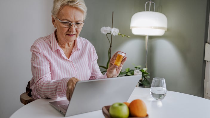 Senior woman reviewing information about her prescription on the computer. She is holding the orange pill bottle in one hand and pointing at the screen with her other.