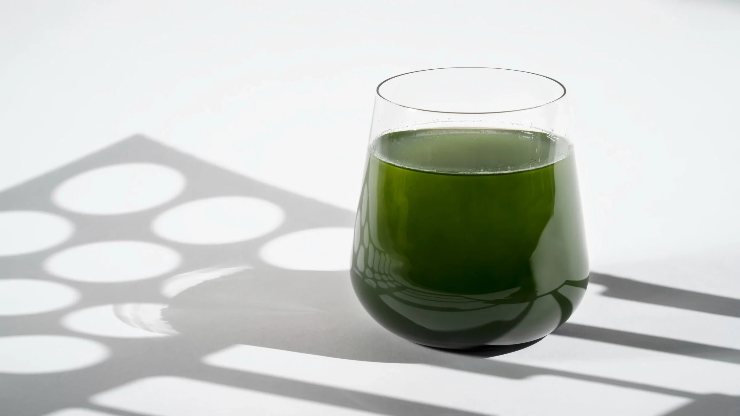 A glass of chlorophyll water on a white background with geometric shadows being cast over it.