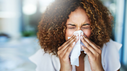 Young woman with curly brown hair blowing her nose. She is wearing a white shirt and the background is blurred.
PeopleImages/iStock via Getty Images