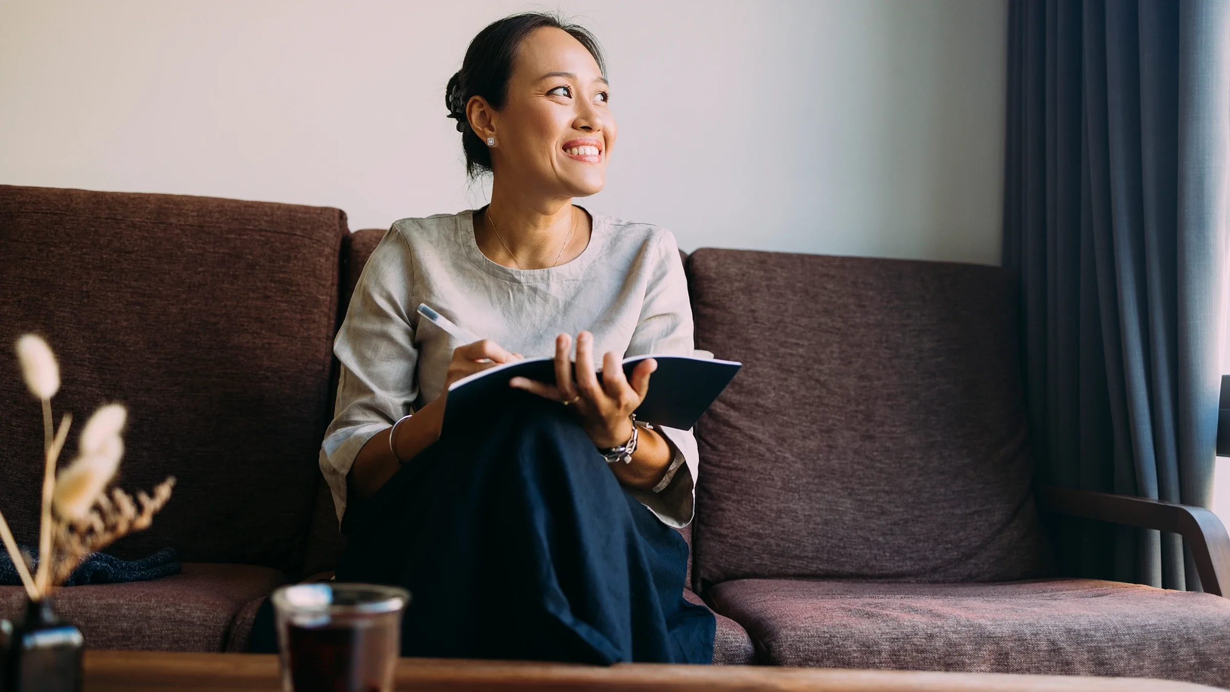 A woman sits comfortably on the couch in her living room, writing in her notebook.