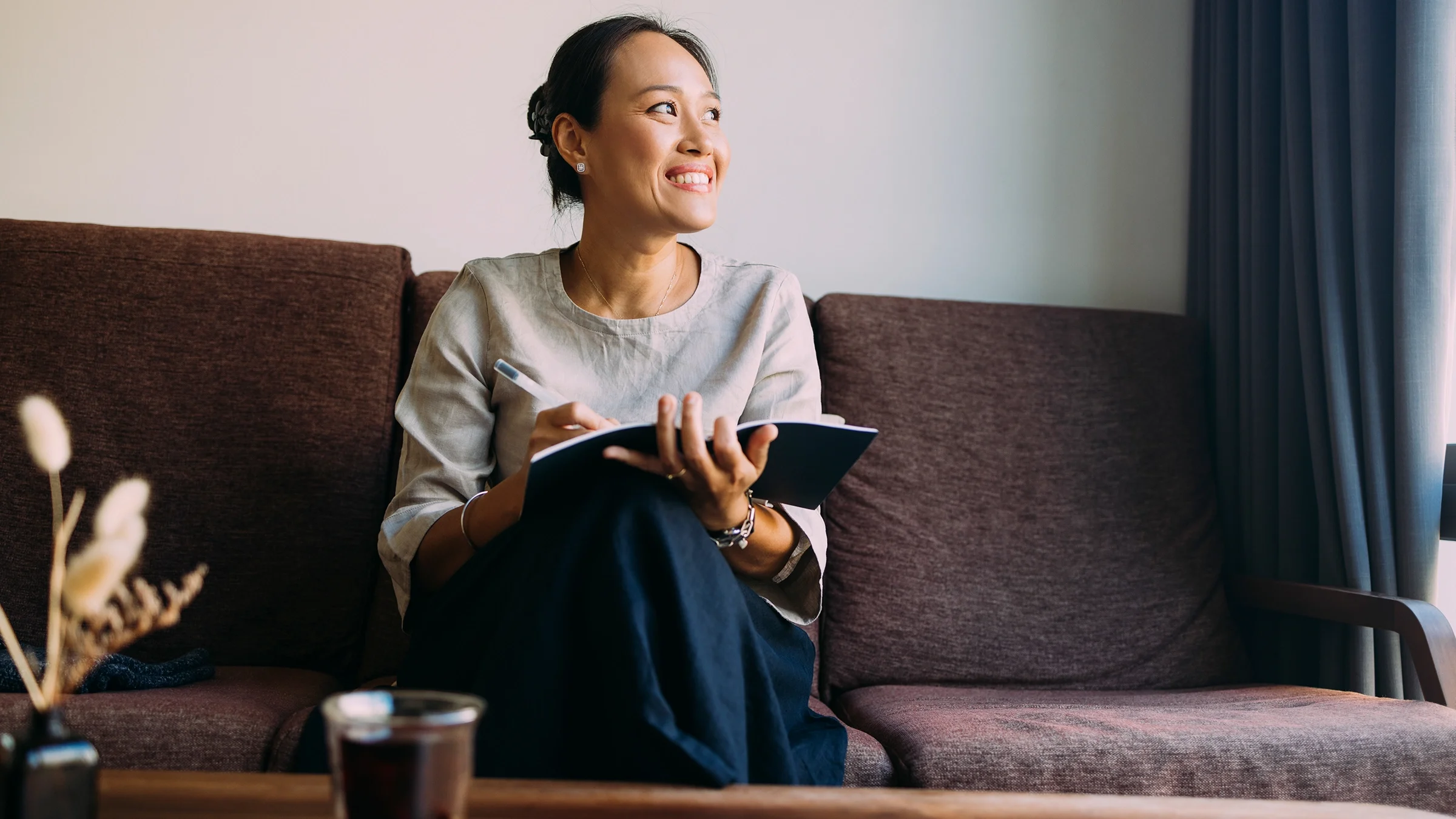 A woman sits comfortably on the couch in her living room, writing in her notebook.