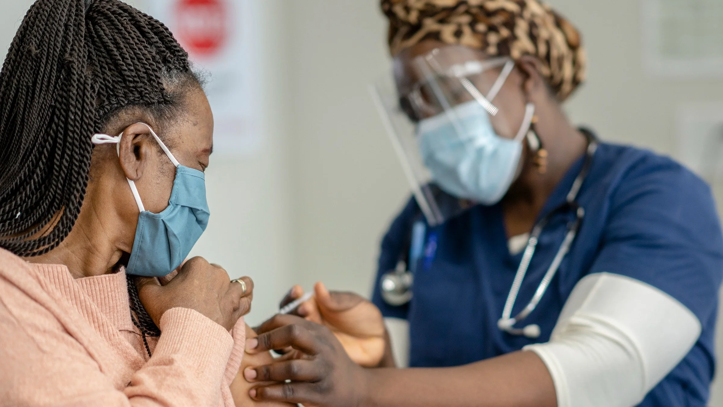 Woman with twists in her hair getting a vaccine from nurse. Both are wearing face masks.