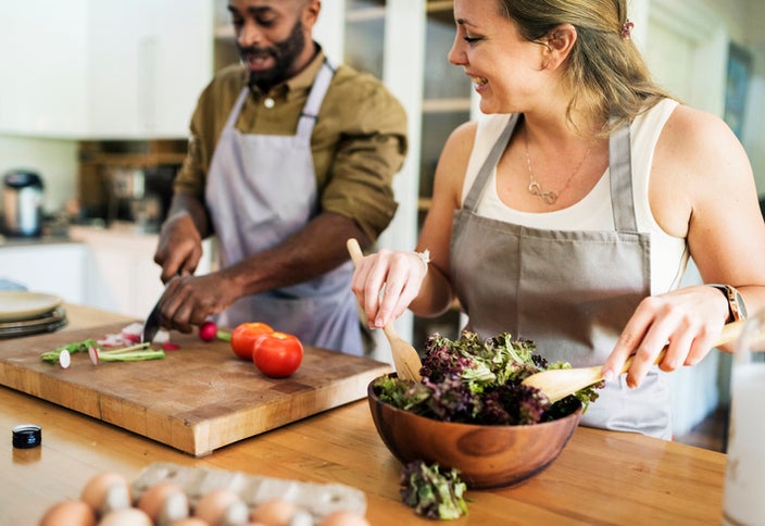 A couple preparing a salad together in the kitchen wearing neutral colored aprons.