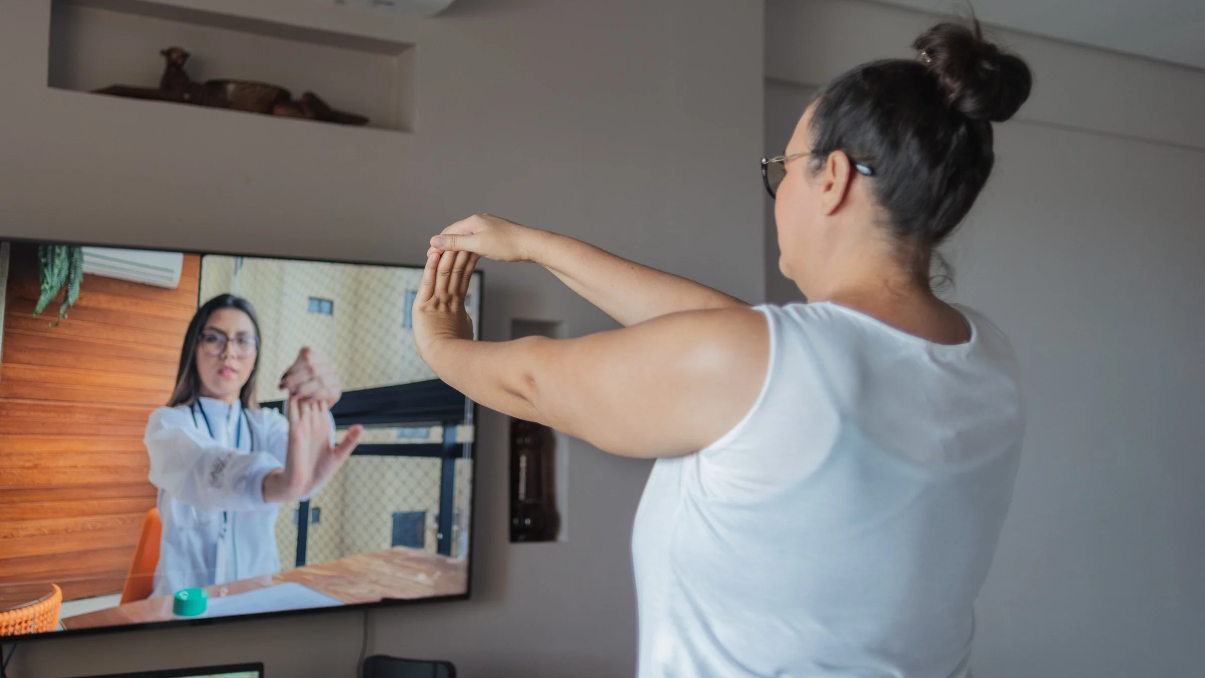 A woman with her hair in a bun doing arm stretches with her doctor on a telehealth visit displayed on her TV.