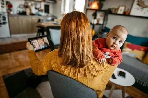 A mother holding her baby on over her shoulder and holding a tablet device in the other hand talking to a nurse on a telehealth visit. The baby is in focus and looking at the camera.
hobo_018/E+ via Getty Images