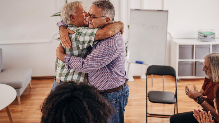 Two men hugging in the middle of a support group circle. They are both smiling.