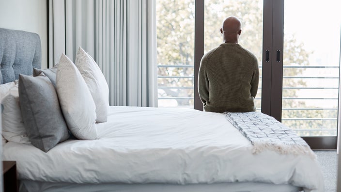 Man sitting on the edge of the bed with his back to the camera. He is looking out the bedroom window.