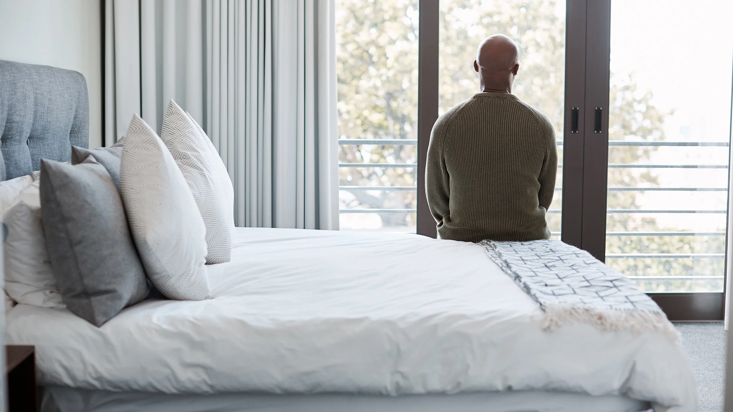 Man sitting on the edge of the bed with his back to the camera. He is looking out the bedroom window.