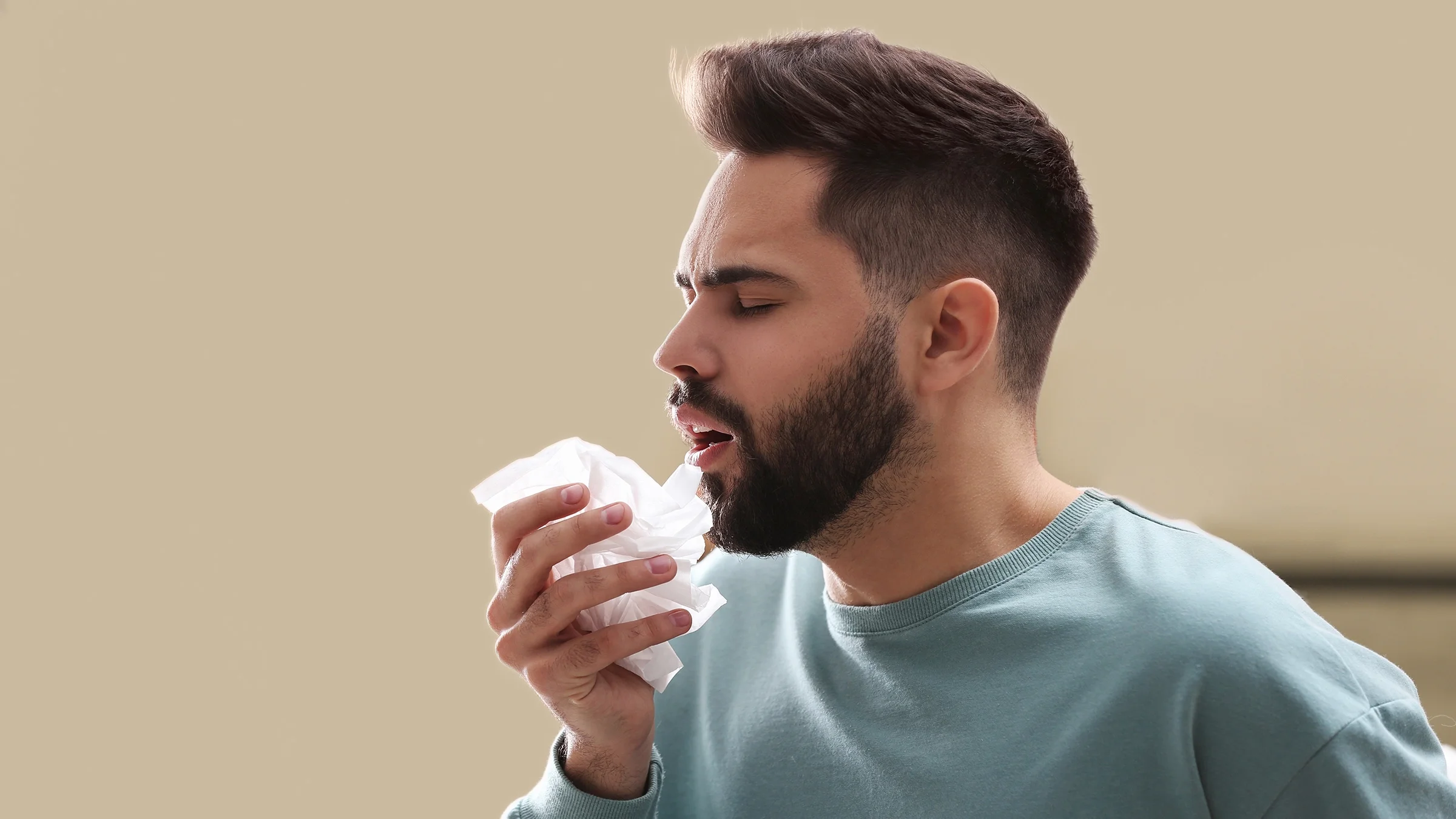 Man sneezing with tissue in hand on a tan background.