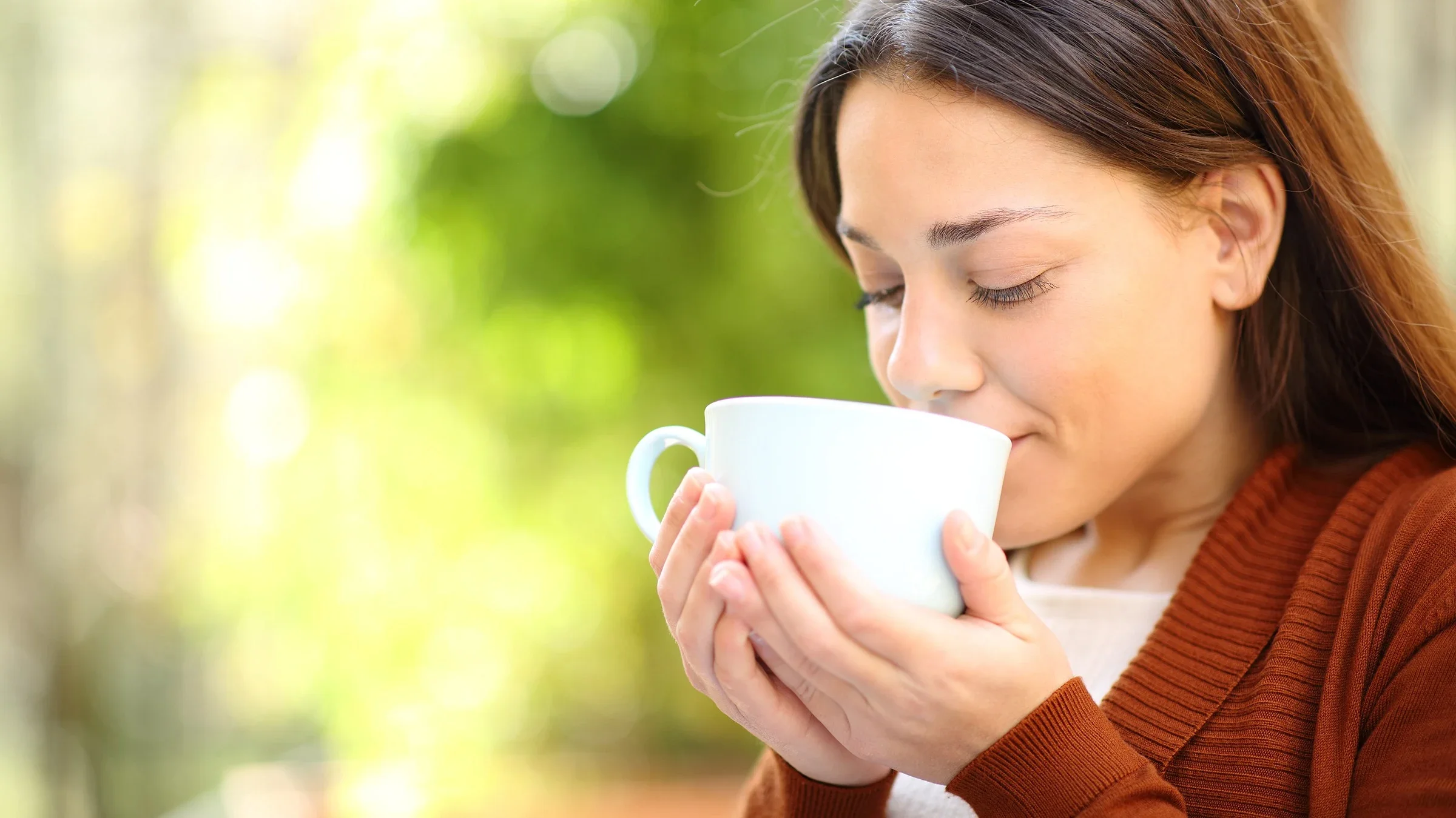 Close-up on a woman drinking a cup of tea. She is smelling and looking down at the white cup with a lush green background behind her.
AntonioGuillem/iStock via Getty Images