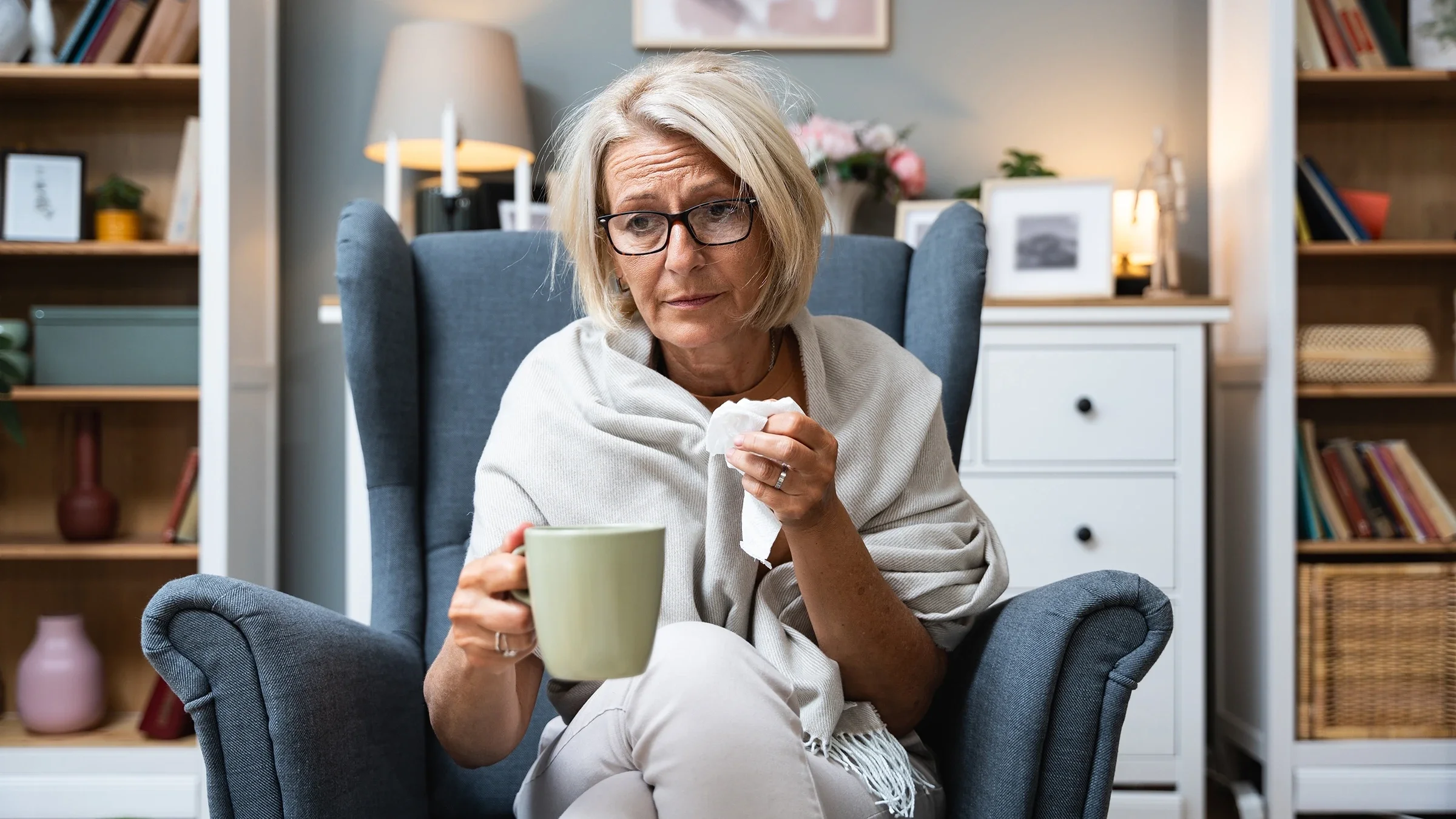 A woman holds a cup of tea and a tissue while sitting on a chair.