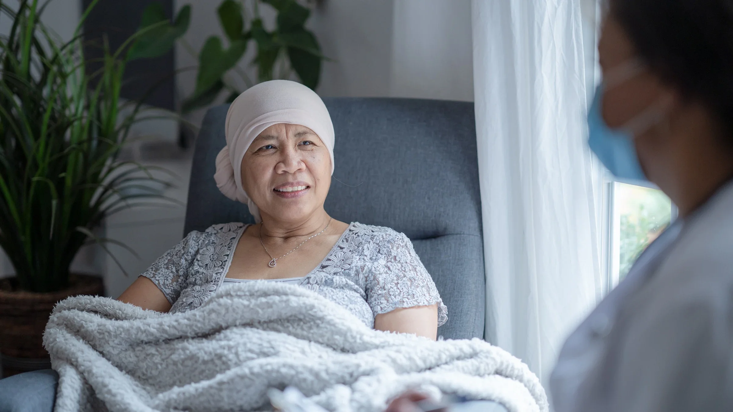 Older woman who is a cancer patient sitting in a chair with a blanket. A doctor is there doing a home visit. They are off to the right of the frame out of focus and cut off on the edge.
FatCamera/E+ via Getty Images
