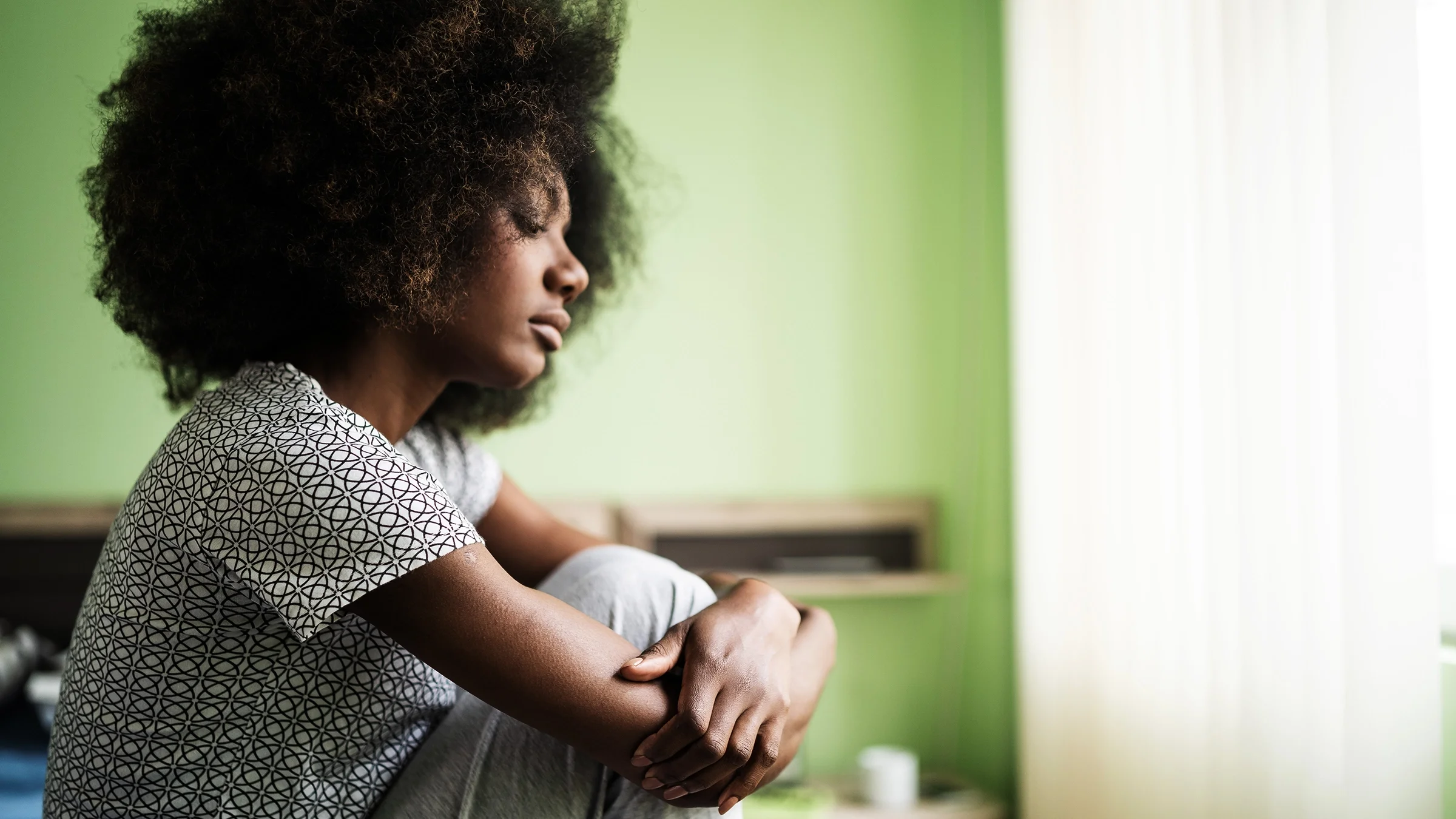 A woman sits on her bed, holding her legs. Having lupus can increase the risk of having mental health conditions like anxiety and depression.