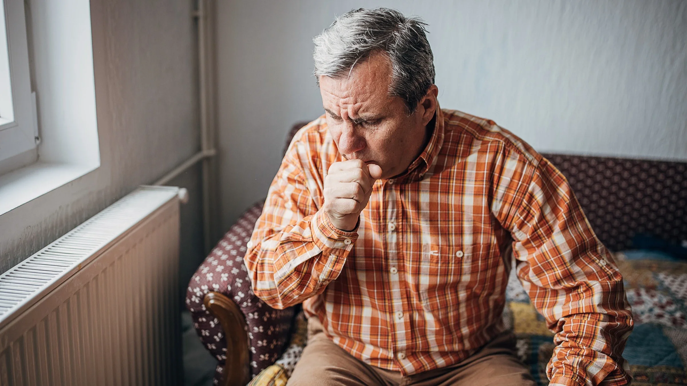 An older adult is coughing. He is sitting on the couch in his home next to the radiator near the window.